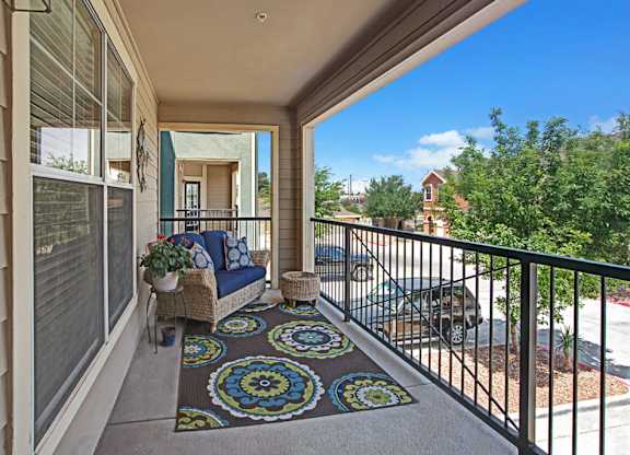 A welcoming private balcony here at Palms at Briarwood featuring comfortable seating, decorative outdoor cushions, and a patterned rug, overlooking landscaped grounds and surrounding neighborhood views while providing an inviting outdoor space to relax and enjoy fresh air.