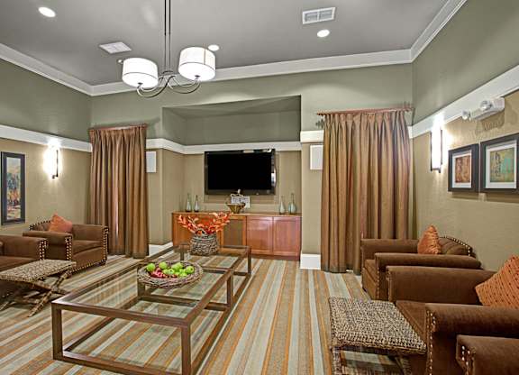 A comfortable resident lounge here at Palms at Briarwood showcasing plush seating, glass coffee tables, a mounted television, and warm neutral tones, with soft lighting, framed wall art, and an inviting layout designed for relaxing, gathering with neighbors, or enjoying casual entertainment.