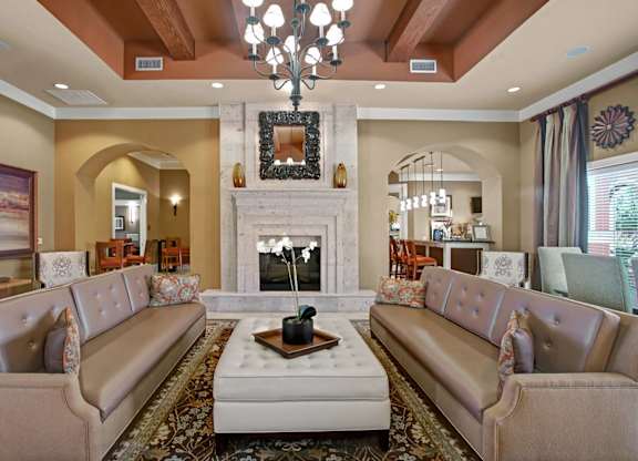 A elegant resident lounge here at Palms at Briarwood featuring symmetrical sofa seating, a large upholstered ottoman, and a statement fireplace with decorative mirror, complemented by a chandelier, exposed ceiling beams, warm neutral tones, patterned area rug, and an inviting layout ideal for relaxing or social gatherings.