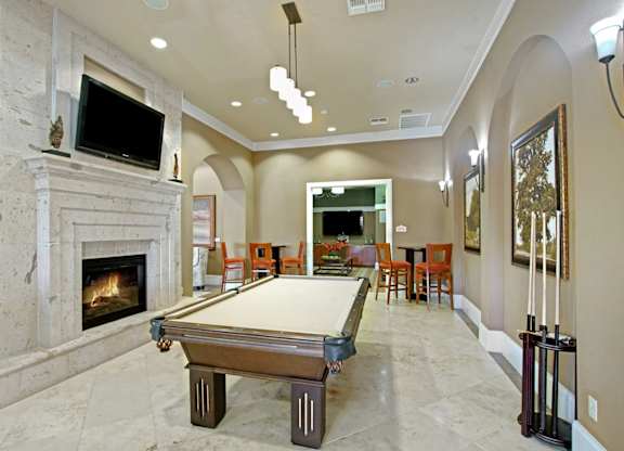 A stylish community game room here at Palms at Briarwood featuring a full-size billiards table, a stone-accent fireplace with mounted television, arched entryways, bar-height seating, pendant lighting, framed artwork, and polished flooring that creates a welcoming space for socializing and recreation.