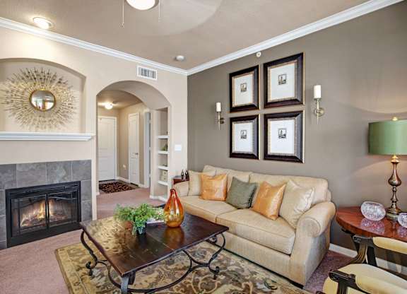 A cozy, well-appointed living room here at Palms at Briarwood featuring plush carpeting, a comfortable sofa with accent pillows, a decorative fireplace with tiled surround, an arched entryway, framed wall art, and a wooden coffee table that creates a warm, inviting space for relaxing or entertaining.