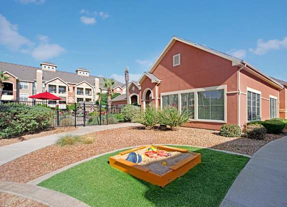 A family-friendly outdoor play area here at Palms at Briarwood featuring a wooden sandbox set on green turf with colorful children’s toys, surrounded by desert-style landscaping, walking paths, manicured shrubs, and nearby community buildings under a bright blue sky.