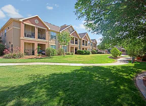 A landscaped apartment exterior here at Palms at Briarwood showcasing multi-story residential buildings with covered balconies, expansive green lawns, mature shade trees, trimmed shrubs, and a curved walking path creating an inviting, park-like community setting under a bright blue sky.