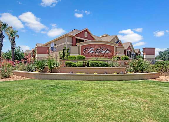 A welcoming community entrance sign here at Palms at Briarwood featuring a monument-style stone sign surrounded by manicured landscaping, ornamental grasses, flowering plants, and trimmed shrubs, with apartment buildings and palm trees in the background beneath a bright blue sky with scattered clouds.
