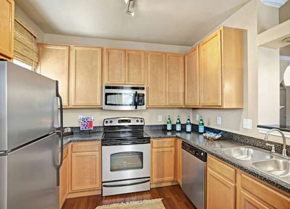 A bright, modern kitchen here at Palms at Briarwood featuring light wood cabinetry, dark stone countertops, a stainless steel refrigerator, oven, microwave, and dishwasher, a double-basin sink, and warm wood-style flooring, with ample cabinet storage and an open layout that connects seamlessly to the adjacent living space.