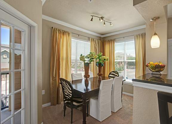 A bright dining area here at Palms at Briarwood featuring a dark wood dining table with seating for four, soft upholstered chairs, and large corner windows that fill the space with natural light, complemented by warm curtains, neutral walls, and an open layout that connects seamlessly to the kitchen and living areas.