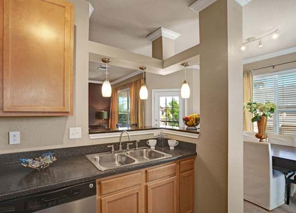 A welcoming kitchen here at Palms at Briarwood featuring warm wood cabinetry, a double-basin sink with a sleek faucet, dark countertops, and pendant lighting above the pass-through, offering an open view into the dining and living areas that creates a bright, connected layout ideal for everyday living and entertaining.