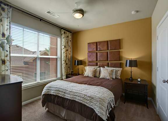 A cozy bedroom here at Palms at Briarwood featuring a comfortable bed with layered bedding, matching nightstands with lamps, soft carpet flooring, and a large window with blinds and patterned curtains that bring in natural light while creating a warm, inviting retreat.
