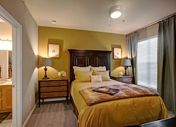 A warm, inviting bedroom here at Palms at Briarwood featuring a dark wood bed frame with layered neutral bedding, matching nightstands with lamps, soft carpeted flooring, and a window with curtains that allows natural light to brighten the cozy, well-appointed space.