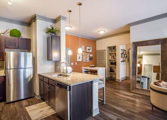 A kitchen and open living area here at Sorrel Fairview with a granite island, dark wood cabinets, stainless steel appliances, warm accent wall, large floor mirror, and a beige chair with brown trim on wood flooring.