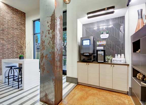 A coffee bar area here at Sorrel Fairview with a self-serve coffee machine, light wood cabinets, dark tile backsplash, brick accent wall, tall metal column, and a white high-top table with black stools.