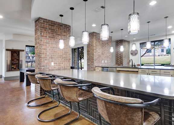 A clubhouse bar and kitchen space here at Sorrel Fairview with a long stone counter, brown leather bar chairs, brick columns, black tile bar base, and modern pendant lights, bright and welcoming.