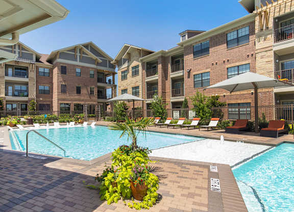 A resort-style pool courtyard here at Sorrel Fairview with a bright blue pool, shallow sun shelf, tan paver deck, and orange lounge chairs beside modern brick apartment buildings.