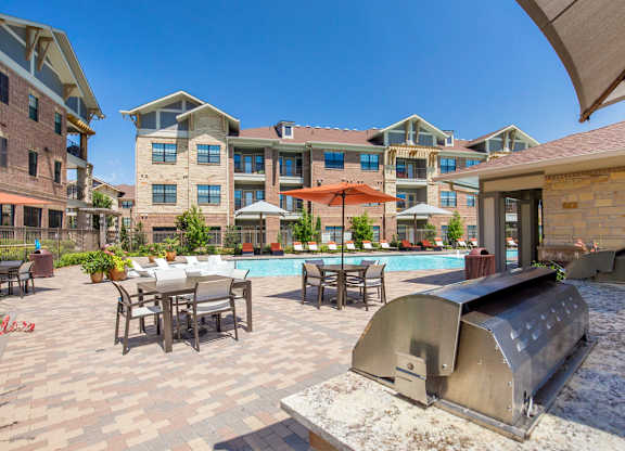 A poolside grilling and dining patio here at Sorrel Fairview with stainless steel grill, stone counter, paver deck, shaded tables with umbrellas, and a bright resort-style pool surrounded by modern brick apartment buildings.