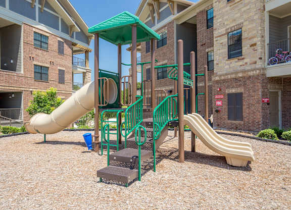 A playground area here at Sorrel Fairview with green and tan climbing structure, twin slides, covered platform, and wood-chip ground, surrounded by brick apartment buildings and sunny blue sky.
