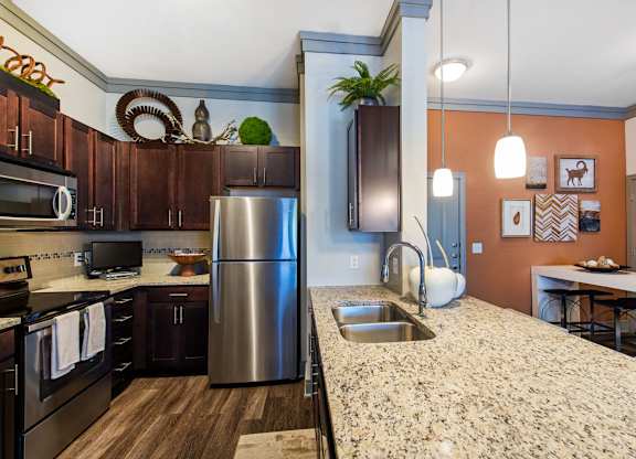 A kitchen here at Sorrel Fairview with a speckled granite island, dark espresso cabinets, stainless steel appliances, pendant lights, and a warm orange accent wall with framed art beside a small white dining nook.