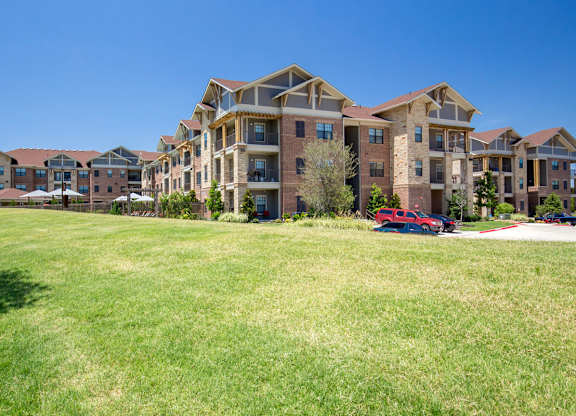 A wide exterior lawn view here at Sorrel Fairview with green open grass leading to multi-story brick and stone apartment buildings with balconies under a clear bright blue sky.
