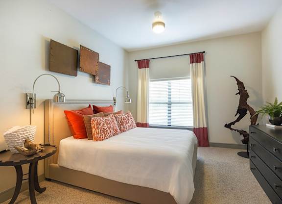A bedroom here at Sorrel Fairview with a beige upholstered bed, white bedding, bold red and patterned accent pillows, black dresser, sculptural wood decor, and cream curtains with red trim over a bright window.