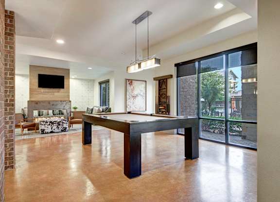 A game lounge here at Sorrel Fairview with a modern pool table, polished concrete floors, brick accent wall, large windows with dark shades, and a cozy seating area with fireplace and TV.