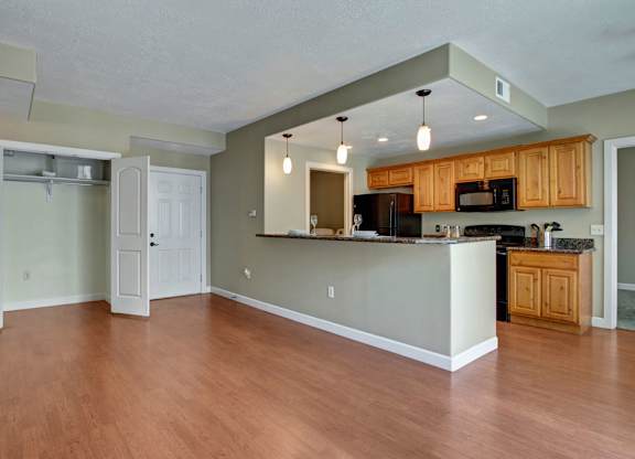 A bright open living and kitchen space here at Bridges with warm wood flooring, natural wood cabinets, a breakfast bar with pendant lights, black appliances, and an open entry closet near the front door.