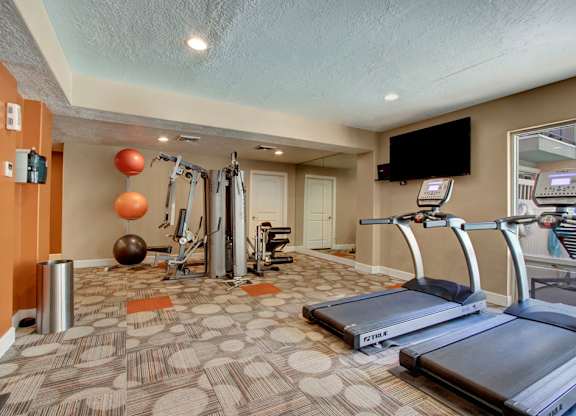 A fitness room here at Bridges with treadmills, a cable weight machine, wall mirrors, mounted TV, patterned carpet, and orange accent walls with exercise balls and strength equipment.