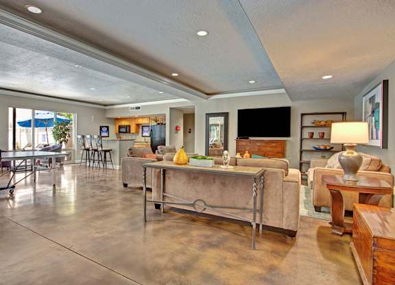 A game and lounge room here at Bridges with tan sofas, wood accent tables, ping pong table, bar-height kitchen counter with stools, TV wall, and polished concrete floors in a bright, open layout.