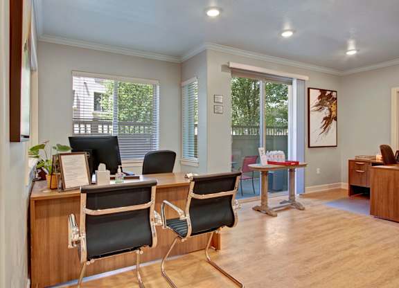 A leasing office here at Bridges with warm wood desks, black guest chairs, light wood floors, and bright windows with patio view, creating a clean, welcoming workspace.