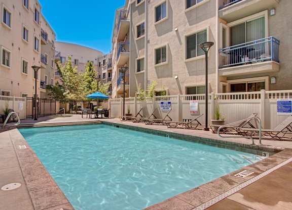 A pool courtyard here at Bridges with a rectangular pool, beige apartment buildings, blue balcony railings, tan deck, and lounge chairs, creating a bright, inviting outdoor amenity space.