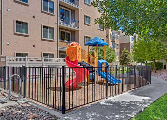 A playground courtyard here at Bridges with a colorful red and blue slide set, black safety fence, green trees, and surrounding apartment balconies, creating a bright, family-friendly outdoor space.