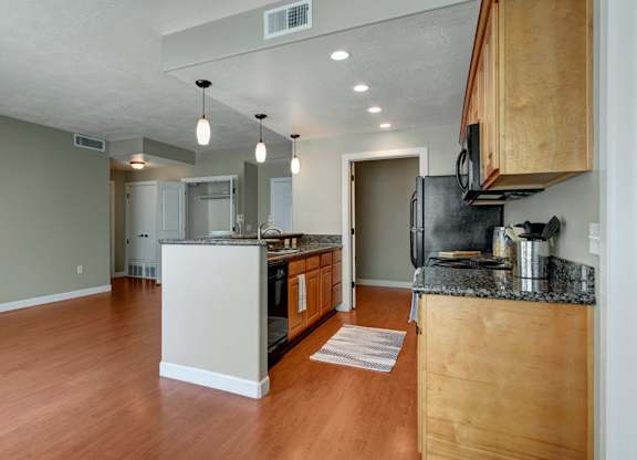 A bright open kitchen and living area here at Bridges with warm wood flooring, light wood cabinets, dark countertops, black appliances, a center island with pendant lights, and an open layout leading to nearby rooms.