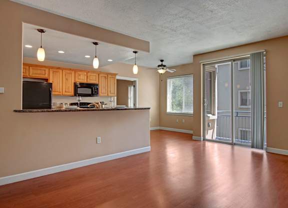 A living and kitchen space here at Bridges with warm wood flooring, light wood cabinets, granite breakfast bar, black appliances, pendant lights, and a bright sliding glass door leading to a private balcony.
