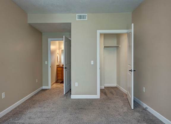 A bedroom here at Bridges with beige walls, gray carpet, an open white closet, and a doorway leading to a bright bathroom vanity area with warm wood cabinetry and mirror lighting.