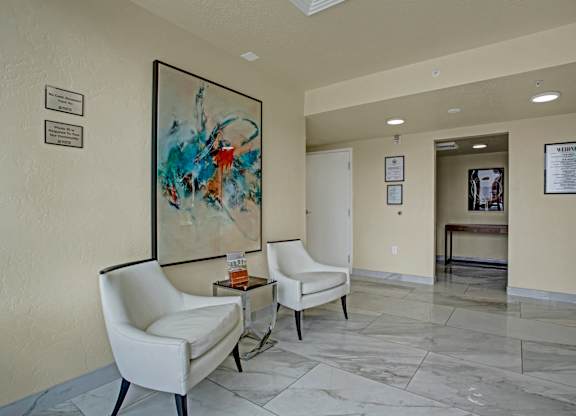 Two white chairs are placed in front of a painting in a hallway.