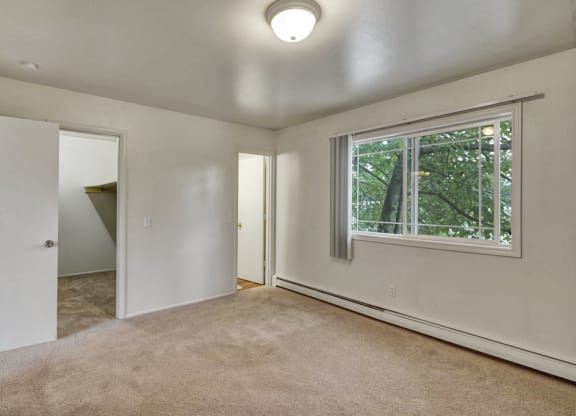 An empty bedroom here at the Village at Calais Apartments with beige carpet, white walls, and a window with vertical blinds showing green foliage outside. Open doors reveal a closet and another room.