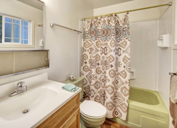 Bright bathroom here at the Village at Calais Apartments with wooden vanity, white sink, and a large mirror. Features a green bathtub, patterned shower curtain, and window providing natural light.
