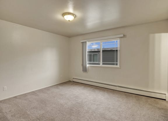 Empty bedroom here at the Village at Calais Apartments with beige carpet and off-white walls, featuring a large window with blinds and a ceiling light. Natural light creates a bright, neutral ambiance.