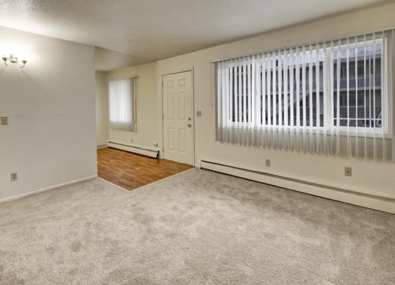 A vacant living room here at Village at Calais Apartments with light beige walls and carpet, featuring a large window with vertical blinds. The entry area has hardwood flooring.
