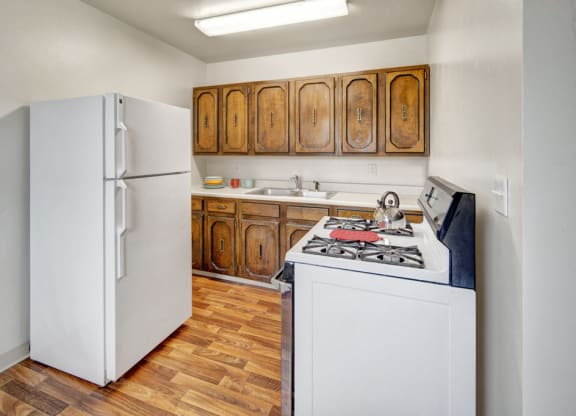 Small kitchen here at the Village at Calais Apartments with wood cabinets, white fridge, and gas stove. Wooden floor, overhead light. Cozy and simple, with a pot on the stove.