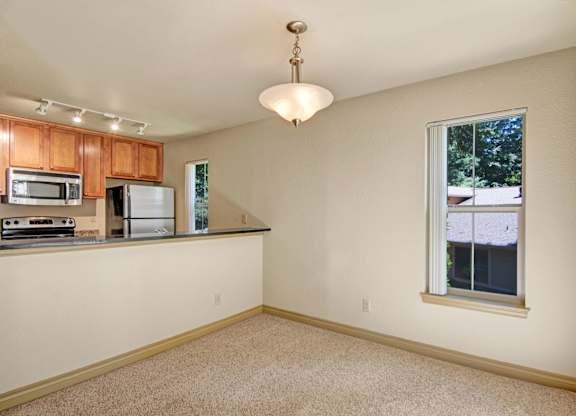 A neutral dining area here at Asbury Park featuring soft carpet flooring, a centered pendant light, and an open pass-through to the kitchen with a dark countertop, warm wood cabinetry, and stainless-steel appliances, while tall windows with vertical blinds bring in natural light and offer views of surrounding greenery.