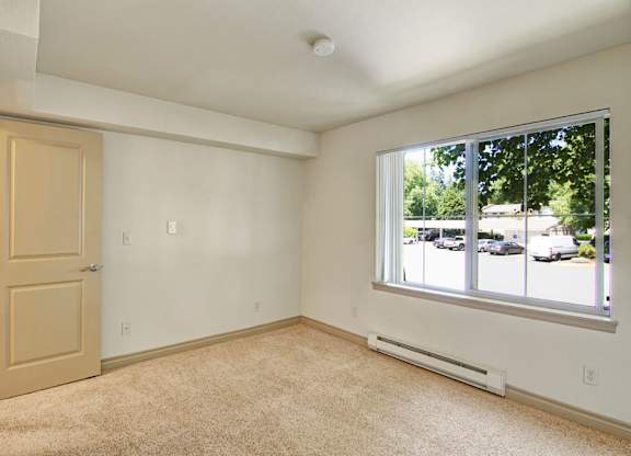 A neutral bedroom here at Asbury Park featuring plush carpet flooring, soft-toned walls, and a wide window with vertical blinds that brings in natural light and offers views of the community parking area, complemented by a simple door entry, clean trim details, and a calm layout ideal for rest and personalization.