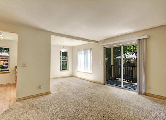 A spacious open living room here at Asbury Park featuring plush carpet flooring, soft neutral walls, and a sliding glass door with vertical blinds that opens to a private balcony, complemented by an adjacent dining nook with a hanging light fixture, large windows for natural light, and a seamless flow into the kitchen area.