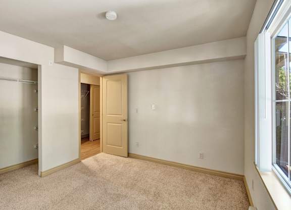 A comfortable bedroom here at Asbury Park featuring soft carpet flooring, neutral-toned walls, and a large window that brings in natural light, complemented by an open closet with shelving, a nearby hallway with wood-style flooring, and ample wall space suitable for a bed, dresser, or additional storage.