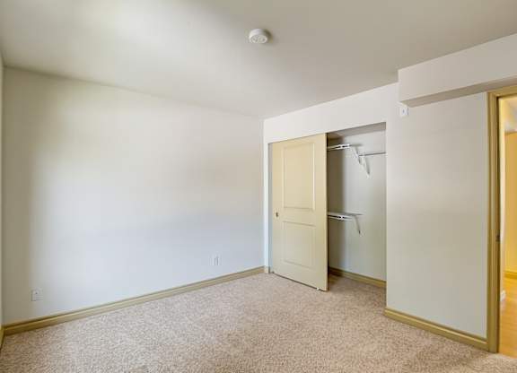 A comfortable bedroom here at Asbury Park featuring plush carpet flooring, soft neutral walls, and a large window that allows natural light to brighten the space, along with an open closet with shelving, a partially open interior door leading to the hallway, and ample wall space suitable for a bed, dresser, or additional storage.