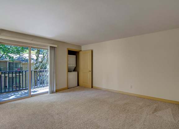 A spacious living room here at Asbury Park featuring plush carpet flooring, soft neutral walls, and a large sliding glass door that opens to a private balcony overlooking mature trees, along with an in-home laundry closet tucked behind a partially open door and an open layout offering ample wall space for seating and entertainment furniture.