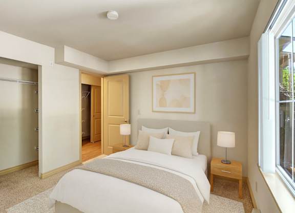 A cozy, well-lit bedroom here at Asbury Park featuring a neatly made bed with white bedding and soft neutral accent pillows, a light upholstered headboard, and matching bedside tables with modern lamps, complemented by plush carpet, an open closet with shelving, and a large window that brings in natural light.