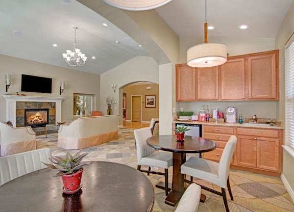 A spacious community lounge here at Asbury Park showcasing a comfortable seating area arranged around a tiled fireplace with a decorative mantel and wall-mounted television, complemented by a kitchenette with wood-style cabinets, countertop appliances, pendant lighting, café-style tables and chairs, soft neutral walls, patterned flooring, and an open layout designed for relaxing and gathering.