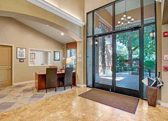 A welcoming leasing office here at Asbury Park featuring a reception desk with guest chairs, a glass entry door framed by floor-to-ceiling windows overlooking landscaped outdoor greenery, tiled flooring, arched architectural details, soft neutral walls, wall signage, and a bright, open layout designed for resident assistance and first impressions.