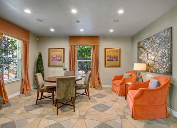 A comfortable resident lounge here at Asbury Park featuring a round dining table with upholstered chairs, two plush accent armchairs with a side table and lamp, patterned carpet flooring, framed wall art, soft neutral walls, ceiling lighting, and large windows with drapery that bring in natural light and create a relaxed gathering space.