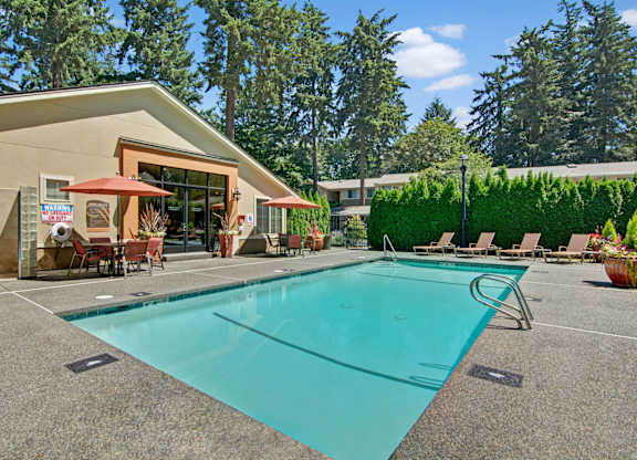 A sunlit outdoor swimming pool here at Asbury Park featuring a clear rectangular pool with metal handrails, a wide textured pool deck, lounge chairs along the edge, shaded patio tables with umbrellas, and a clubhouse building with large windows, all surrounded by tall evergreens and manicured hedges creating a relaxed community setting.