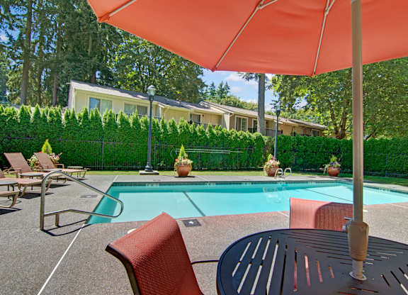 A shaded poolside seating area here at Asbury Park featuring a round outdoor table with chairs beneath a large umbrella, overlooking a clear rectangular swimming pool with metal handrails, surrounding lounge chairs, potted planters, and a fenced perimeter framed by tall evergreen hedges and mature trees in a peaceful community setting.